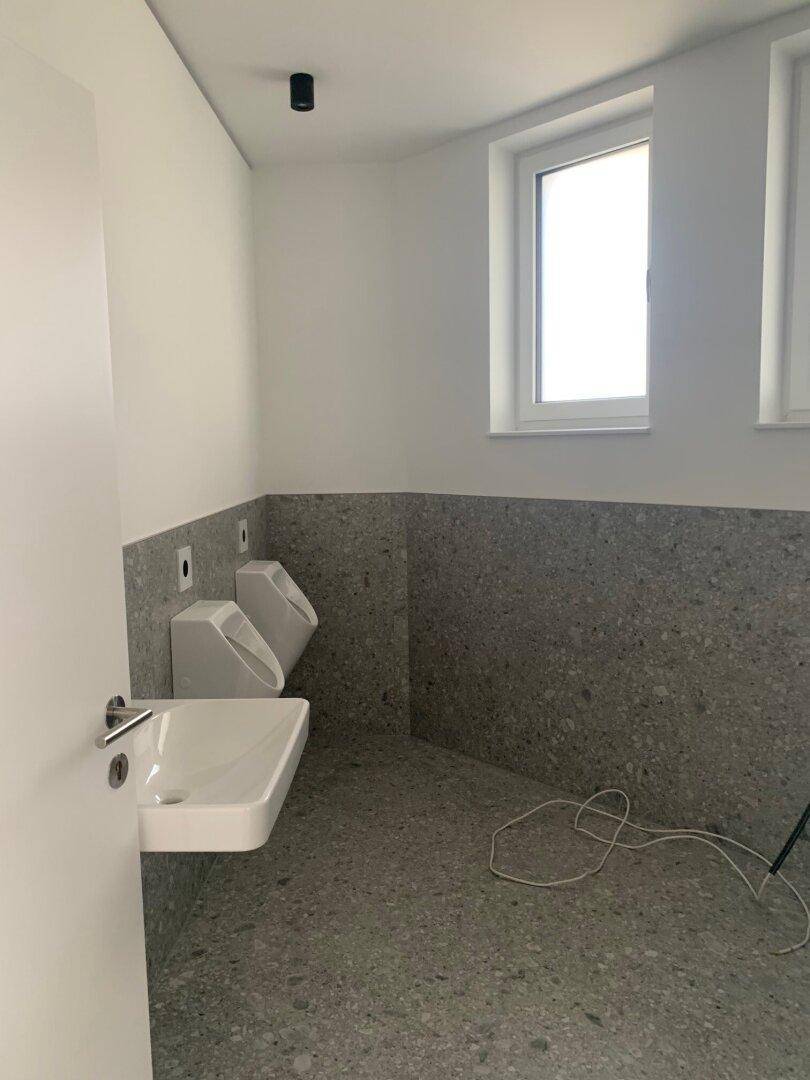 Modernes Badezimmer mit grauen Fliesen, ausgestattet mit Waschbecken und zwei Urinalen.