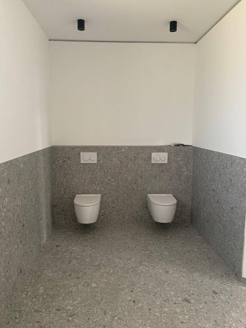 Modernes Badezimmer mit grauen Fliesen und zwei wandmontierten Toiletten.