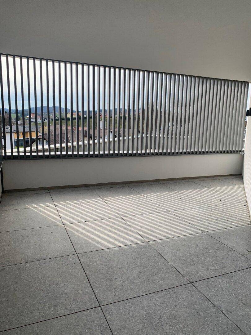 Heller Balkon mit grauen Fliesen und vertikalen Lamellen, der viel Sonnenlicht einfängt.