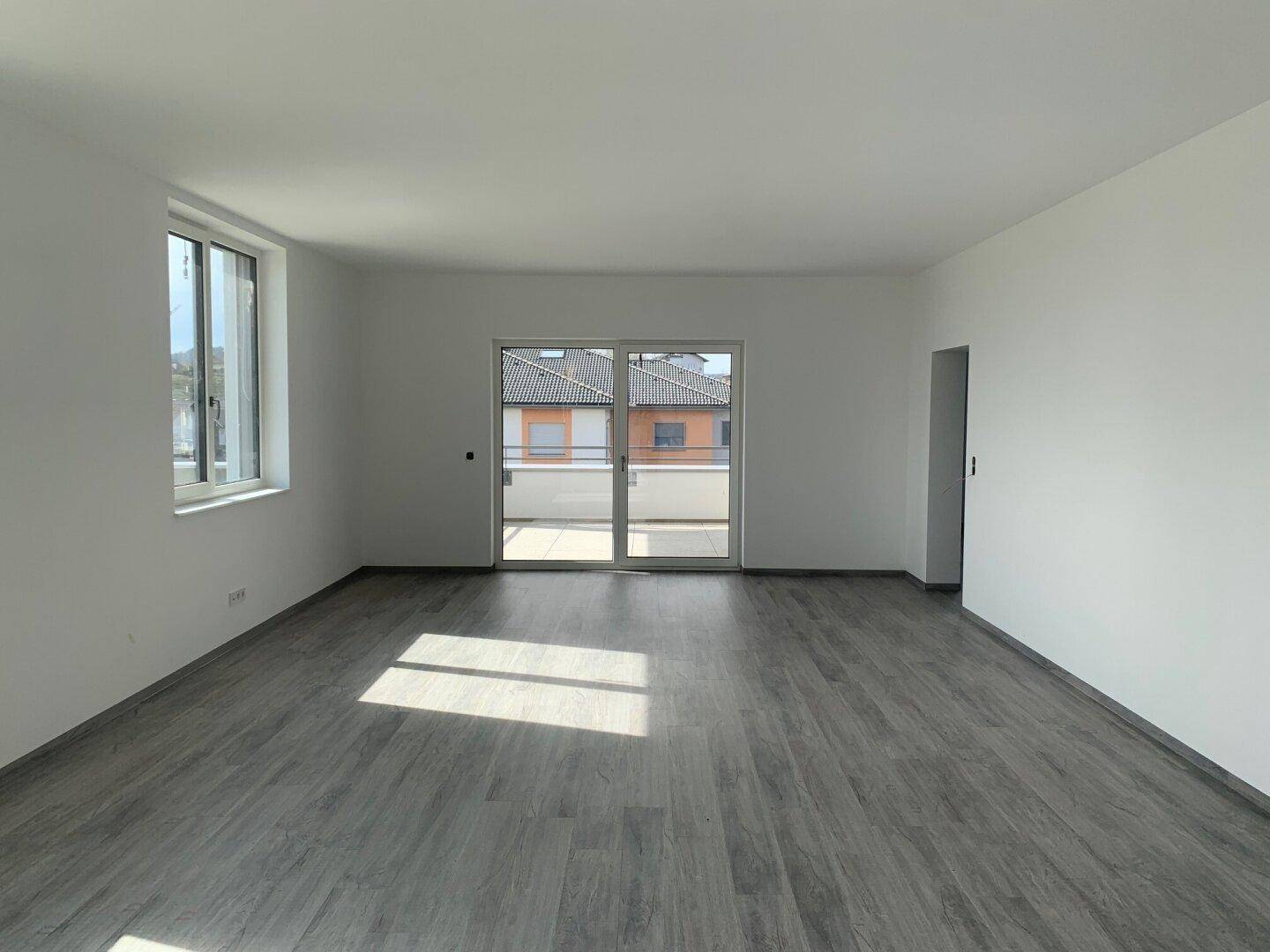 Geräumiger, leerer Raum mit modernem grauen Plankenboden und Zugang zu einem Außenbereich durch eine Glastür.