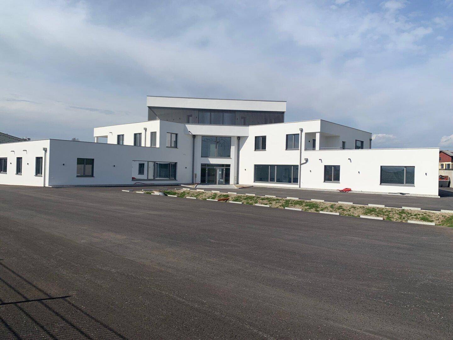 Modernes, weißes Gebäude mit großen Fenstern und weitläufigem Parkplatz, präsentiert zeitgenössische Architektur.