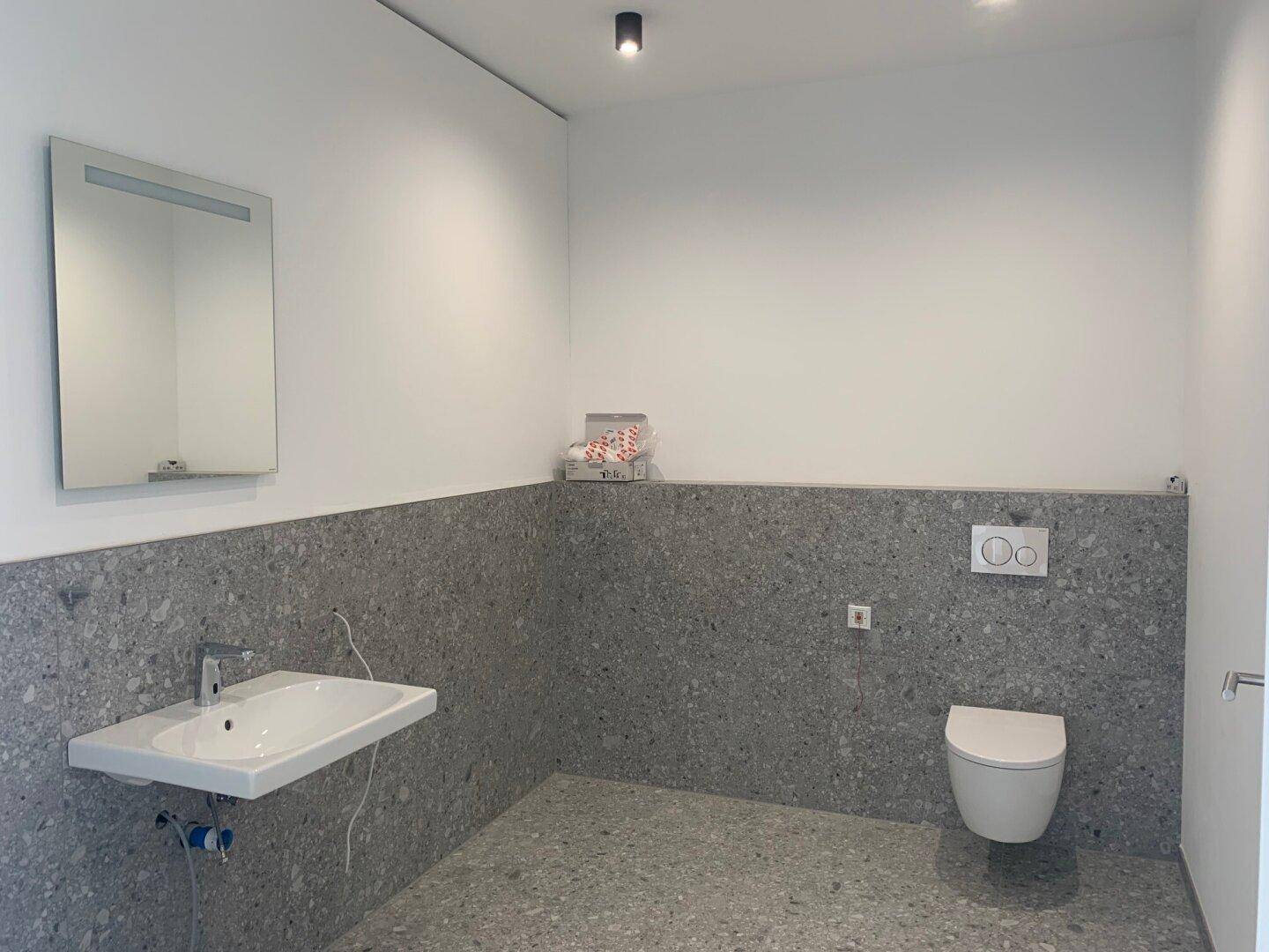 Helles Badezimmer mit modernem Waschbecken, Spiegel und Wand-WC, graue Fliesen an Boden und Wänden.