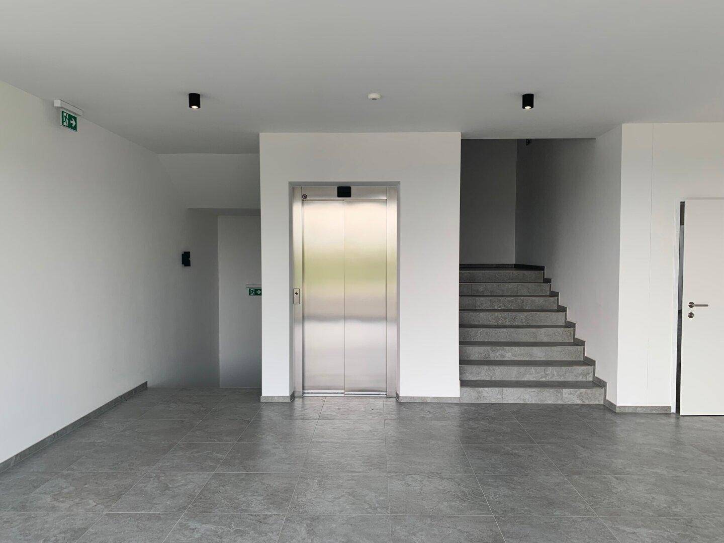 Heller Eingangsbereich mit modernem Aufzug und Treppe, ausgestattet mit grauen Fliesen.