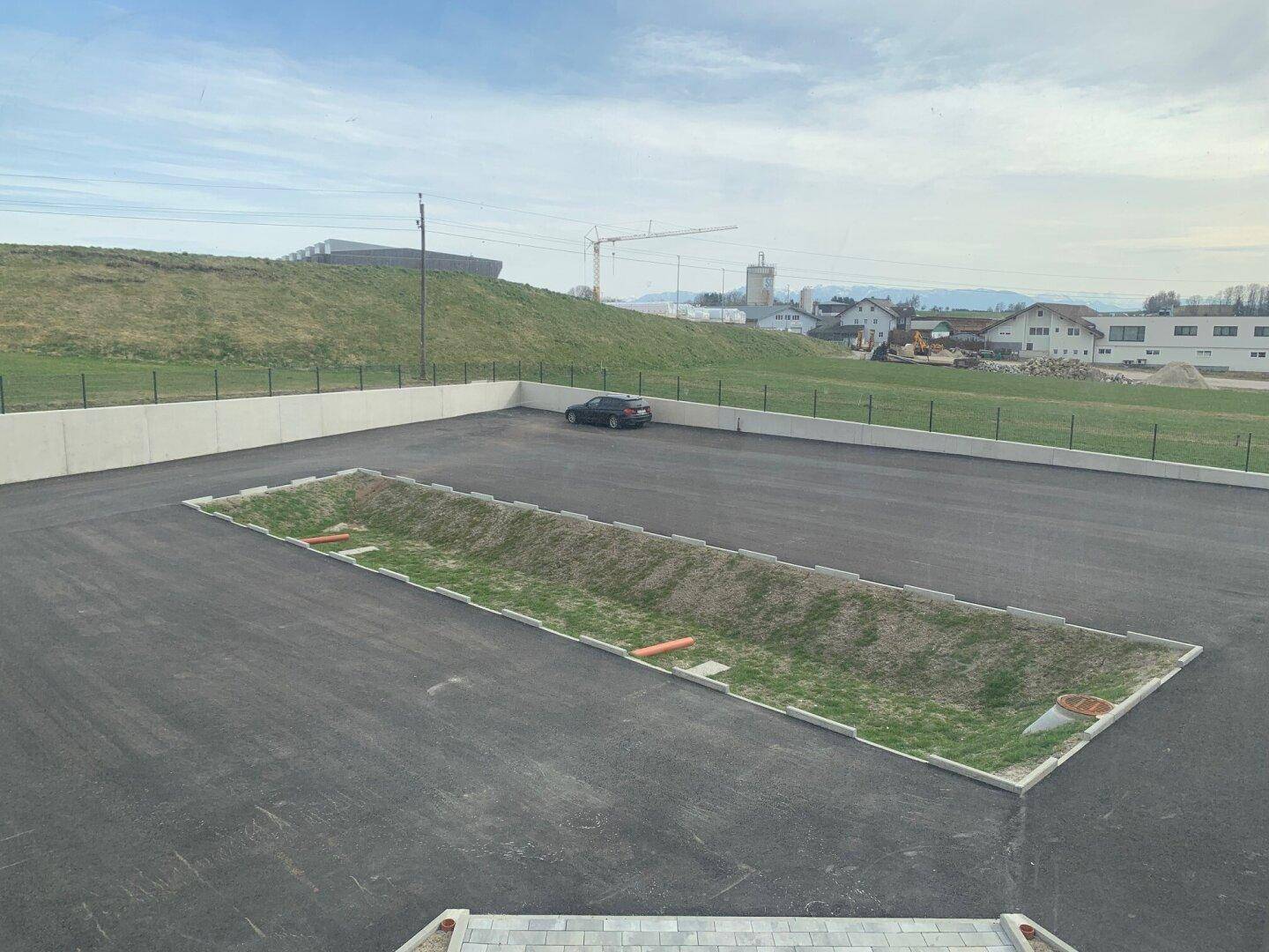 Großer Außenparkplatz mit asphaltierter Fläche und einem begrünten Mittelstreifen, umgeben von Hügeln.