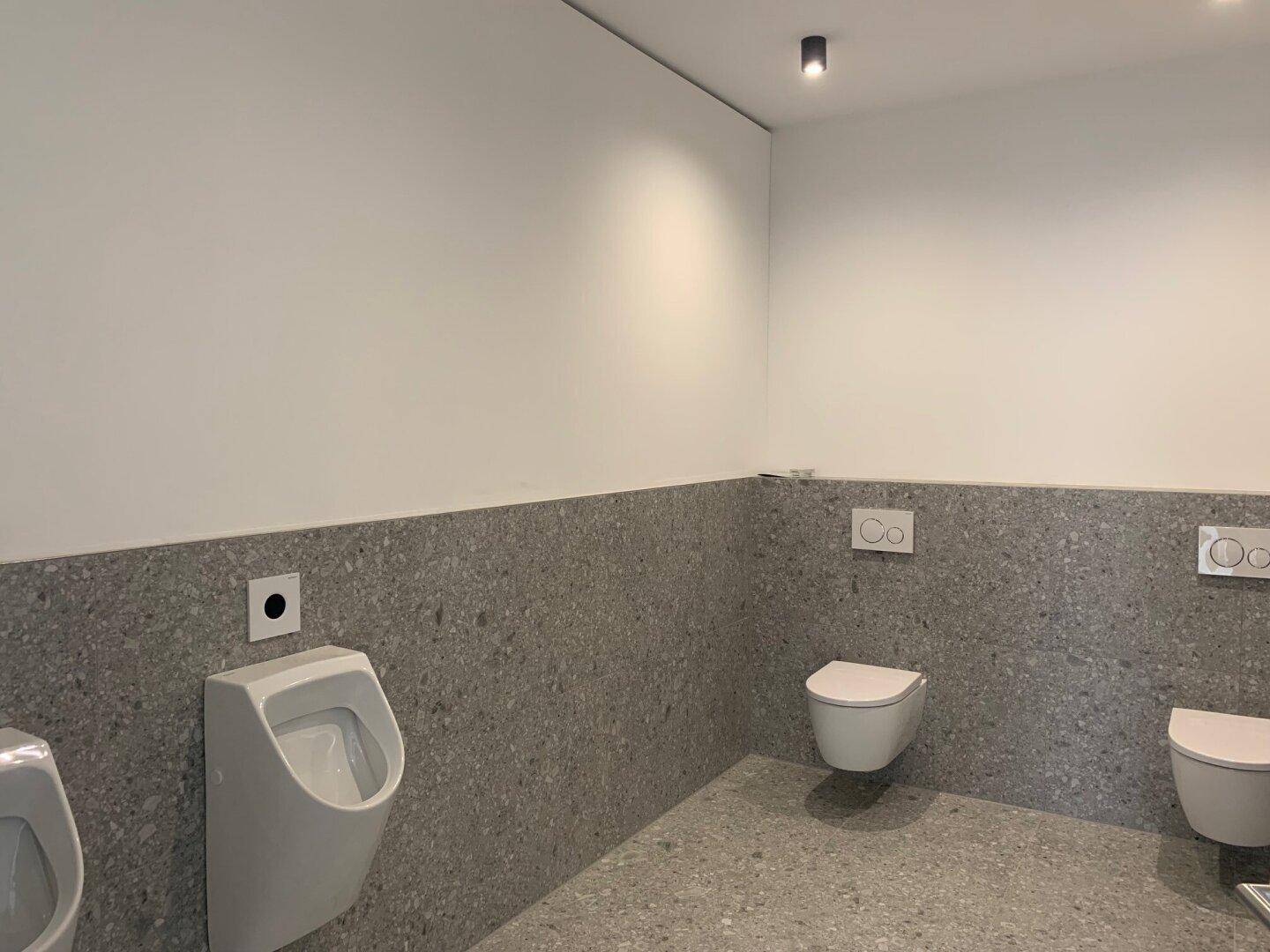 Geräumiges Badezimmer mit mehreren wandhängenden Toiletten und Urinalen, grauen Fliesen und weißen Wänden.