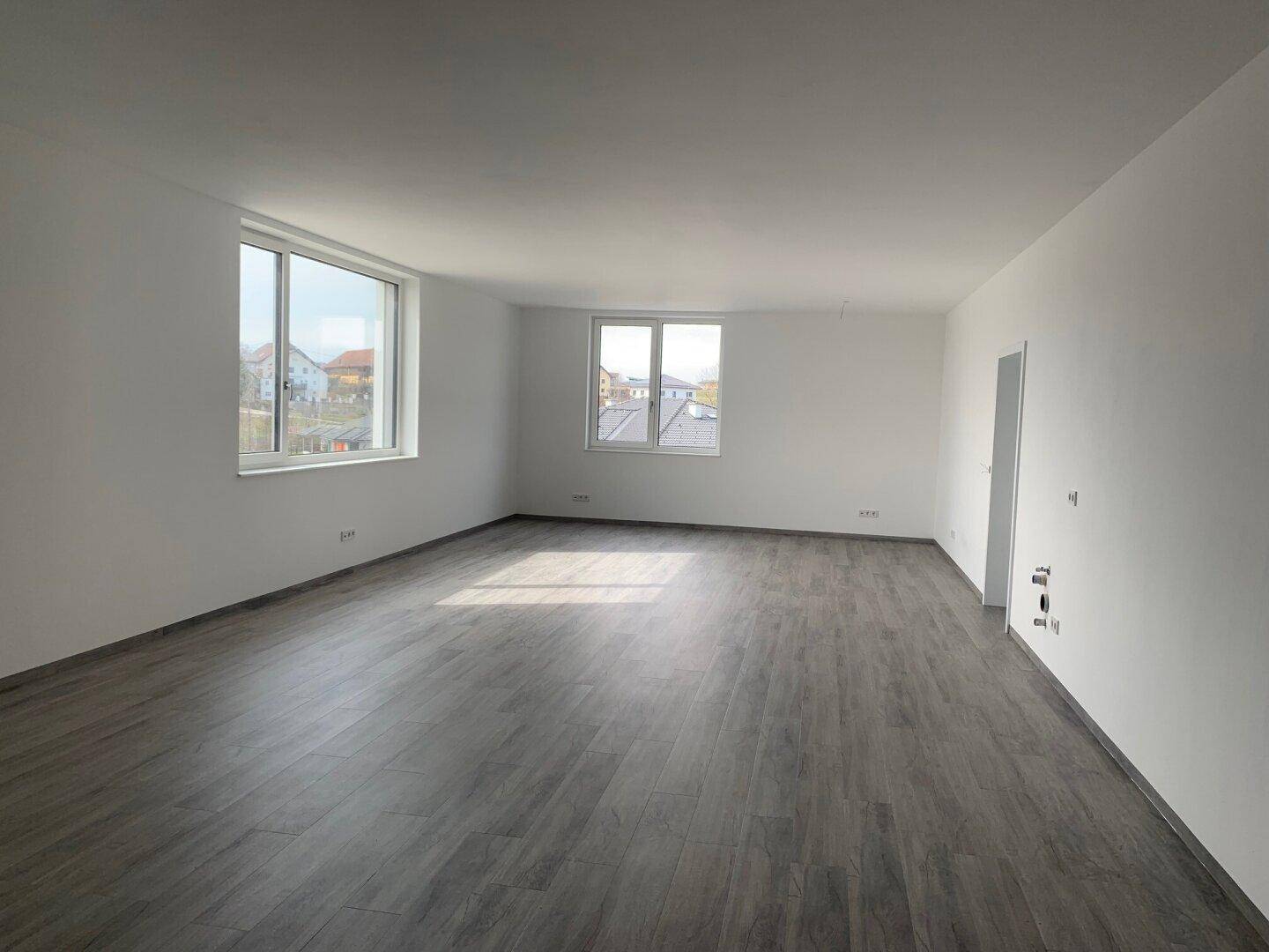 Heller, leerer Raum mit großen Fenstern und modernem grauem Laminatboden, bereit für die Einrichtung.