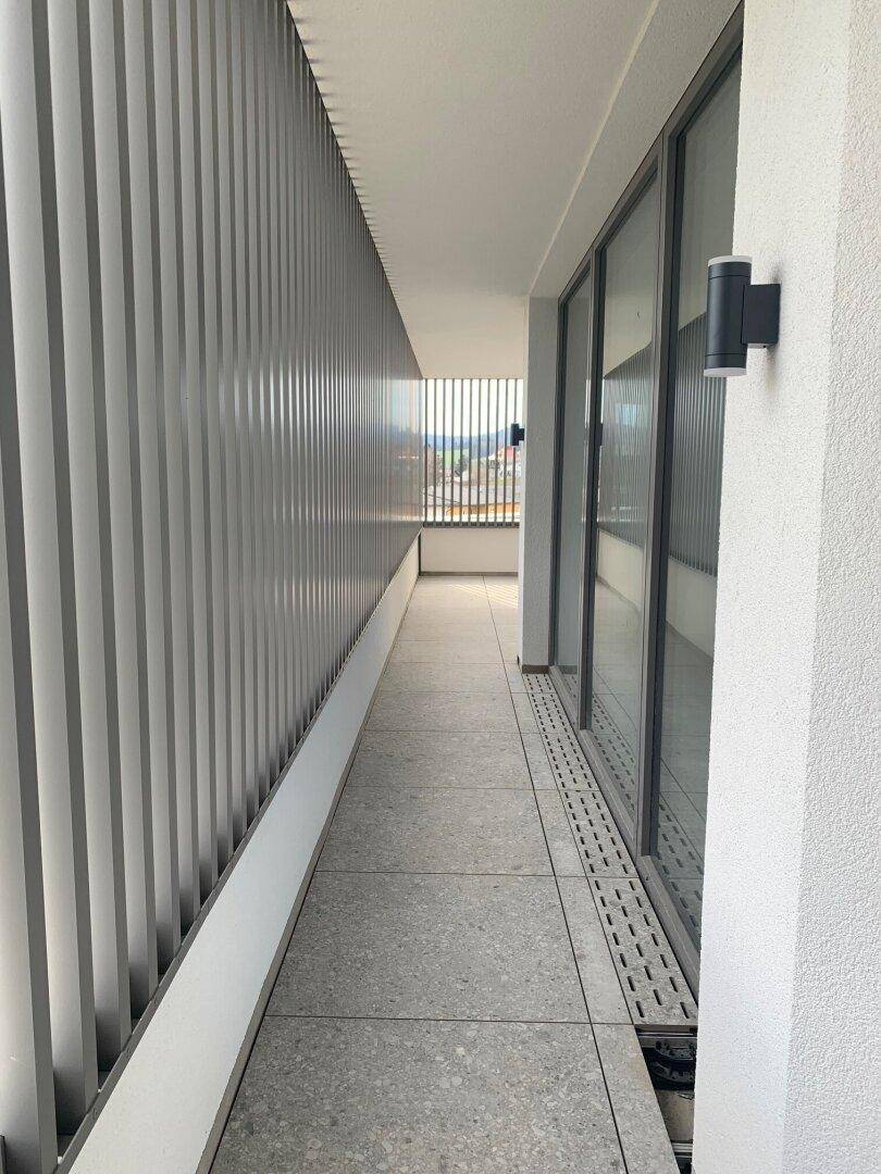 Langer Balkon mit Fliesenboden und Lamellenbrüstung, der entlang der Gebäudeseite verläuft und Ausblick bietet.