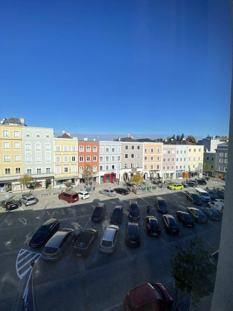 Panoramablick auf einen belebten Stadtplatz mit parkenden Autos und farbenfrohen Gebäuden unter blauem Himmel.