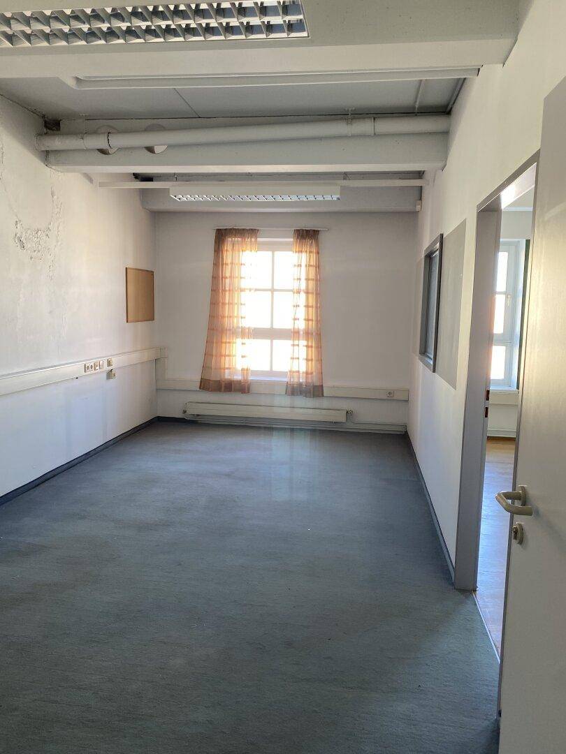 Leeres Büro mit Teppichboden, zwei Fenstern mit Vorhängen und Heizkörpern unter den Fenstern.