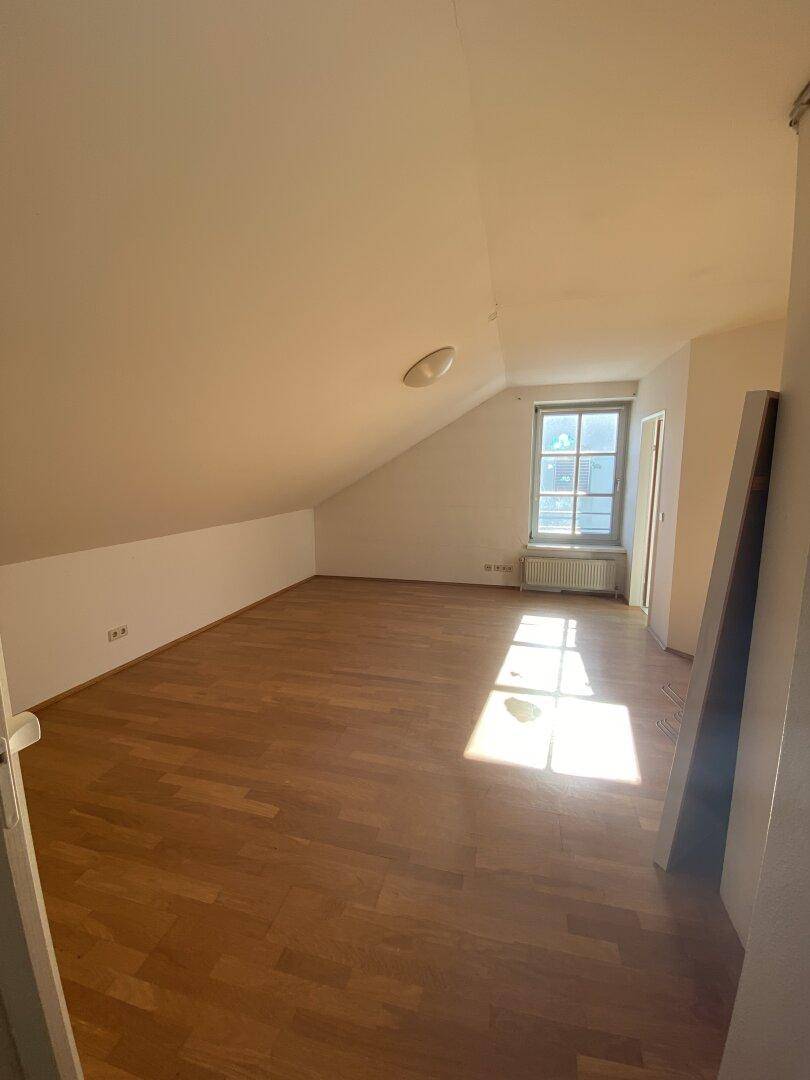 Schlafzimmer im Dachgeschoss mit schrägen Wänden, Holzboden und einem Fenster, das Tageslicht hereinlässt.