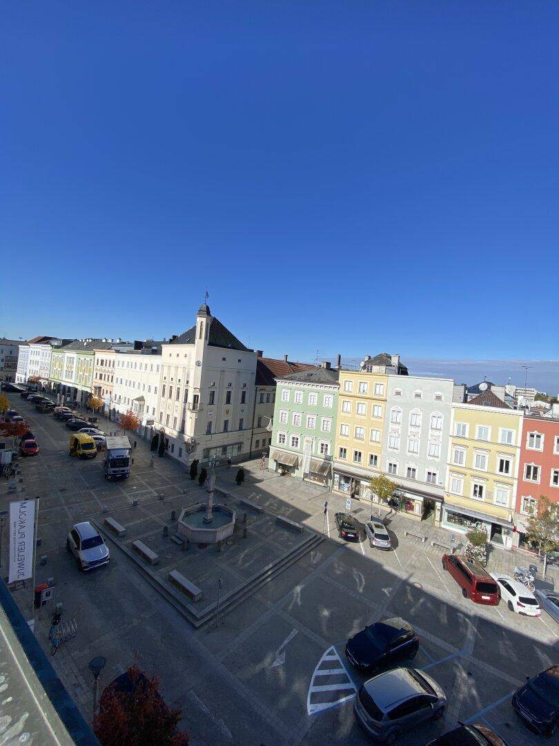 Weitläufiger Blick über einen Stadtplatz mit historischen Gebäuden und parkenden Autos unter blauem Himmel.