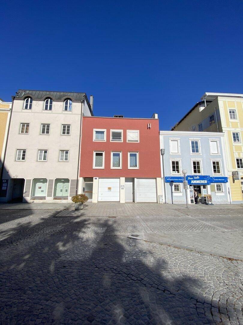 Eine Reihe von farbigen Gebäudefassaden an einem sonnigen Platz mit Kopfsteinpflaster und Garagentoren im Erdgeschoss.
