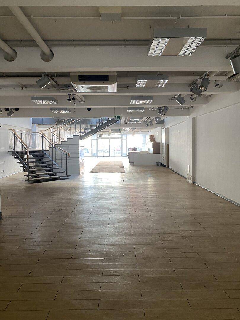 Weitläufiger, leerer Ladenbereich mit Holzboden, einer modernen Treppe und Blick auf die Eingangstür.