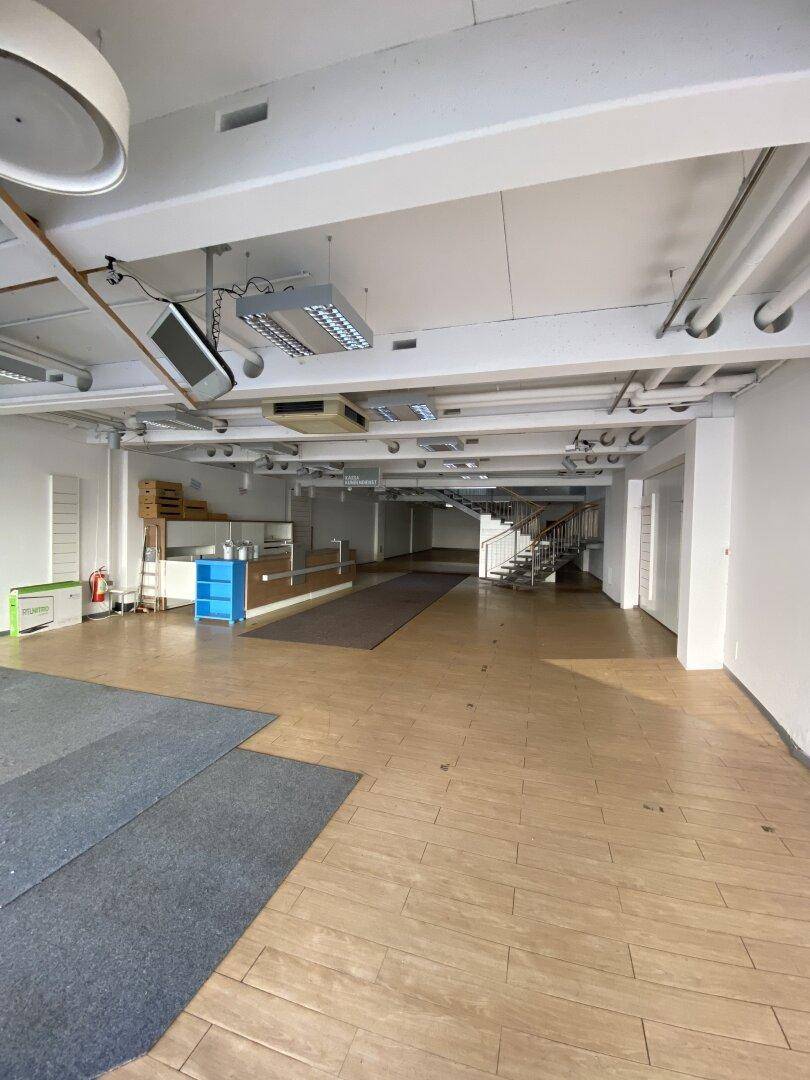 Großer, leerer Ladenbereich mit Holzboden, Teppichabschnitten und einer modernen Treppe im Hintergrund.