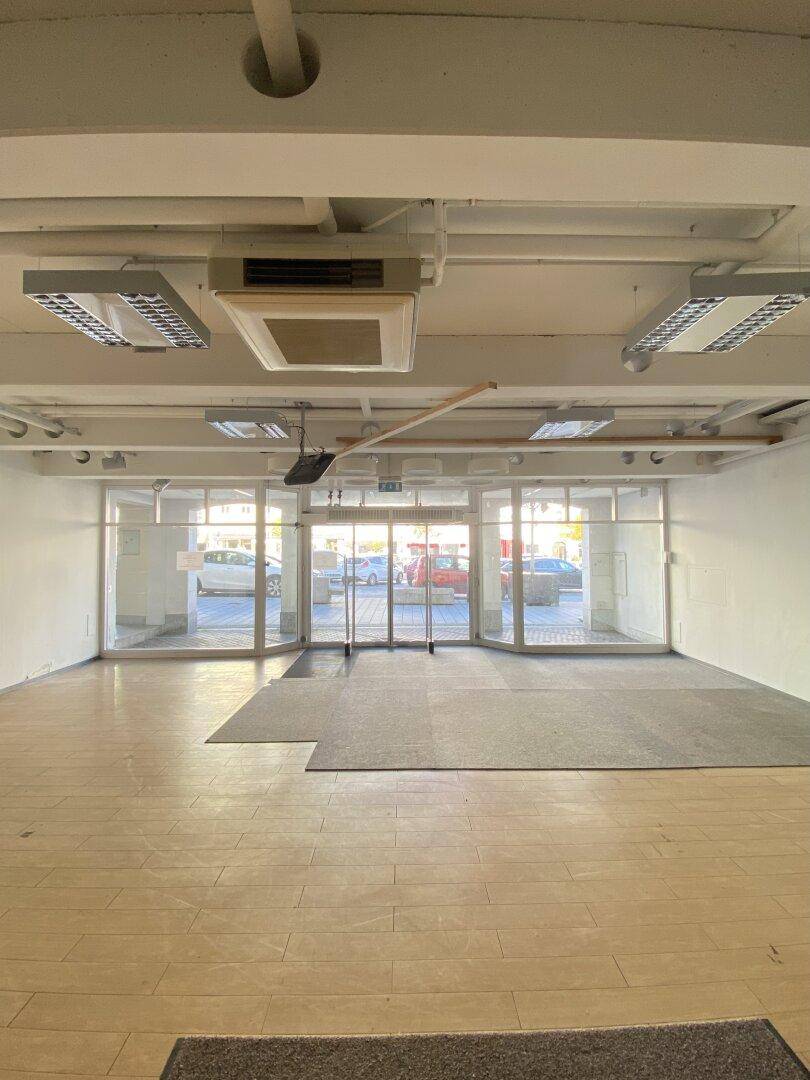 Geräumiger, leerer Ladenbereich mit großen Schaufenstern zur Straße, Holzboden und sichtbaren Deckeninstallationen.