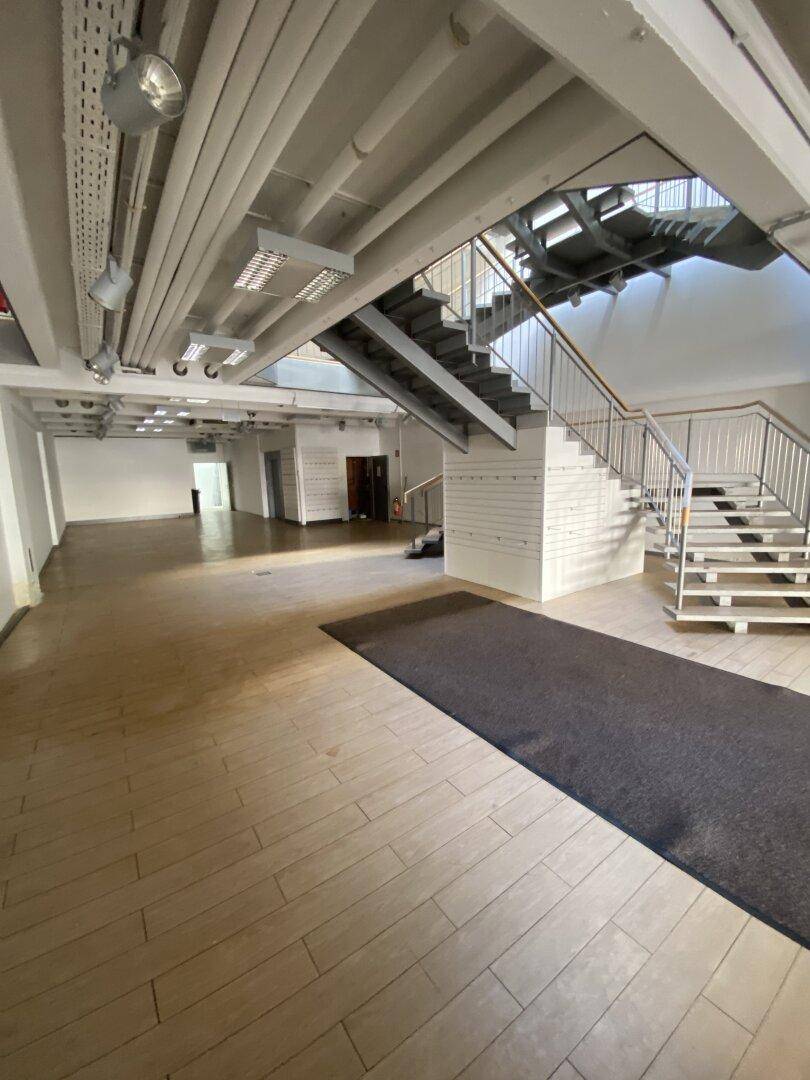 Geräumiger, leerer Ladenbereich mit Holzboden, Teppichabschnitt und einer modernen Treppe im Hintergrund.