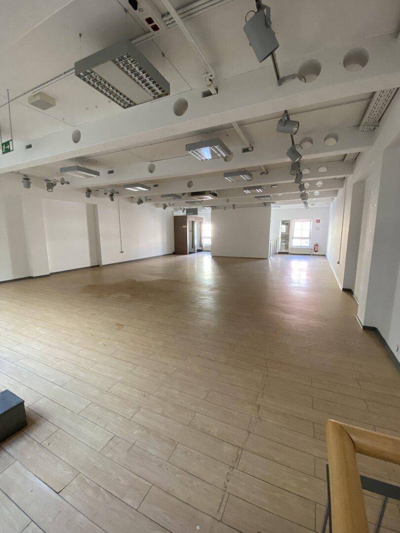 Weitläufiger, leerer Ladenbereich mit Holzboden, weißen Wänden und sichtbaren Deckeninstallationen.