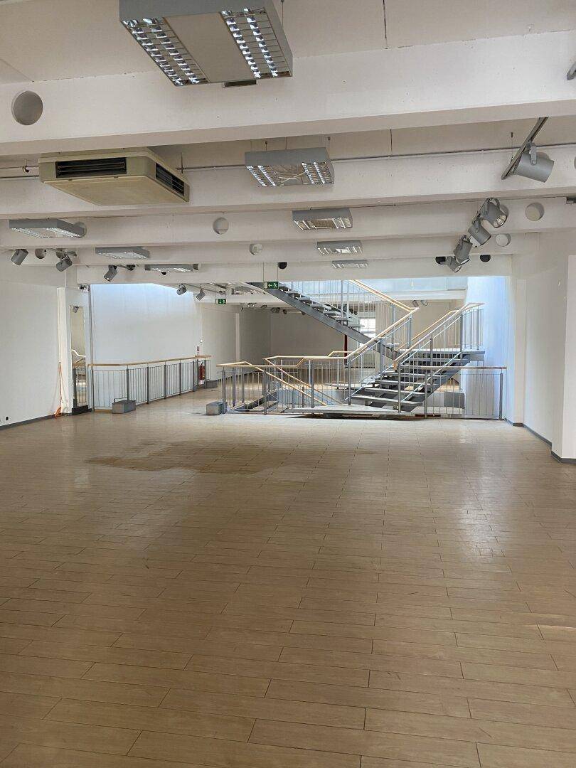 Geräumiger, leerer Ladenbereich mit Holzboden, einer modernen Treppe und sichtbaren Deckeninstallationen.