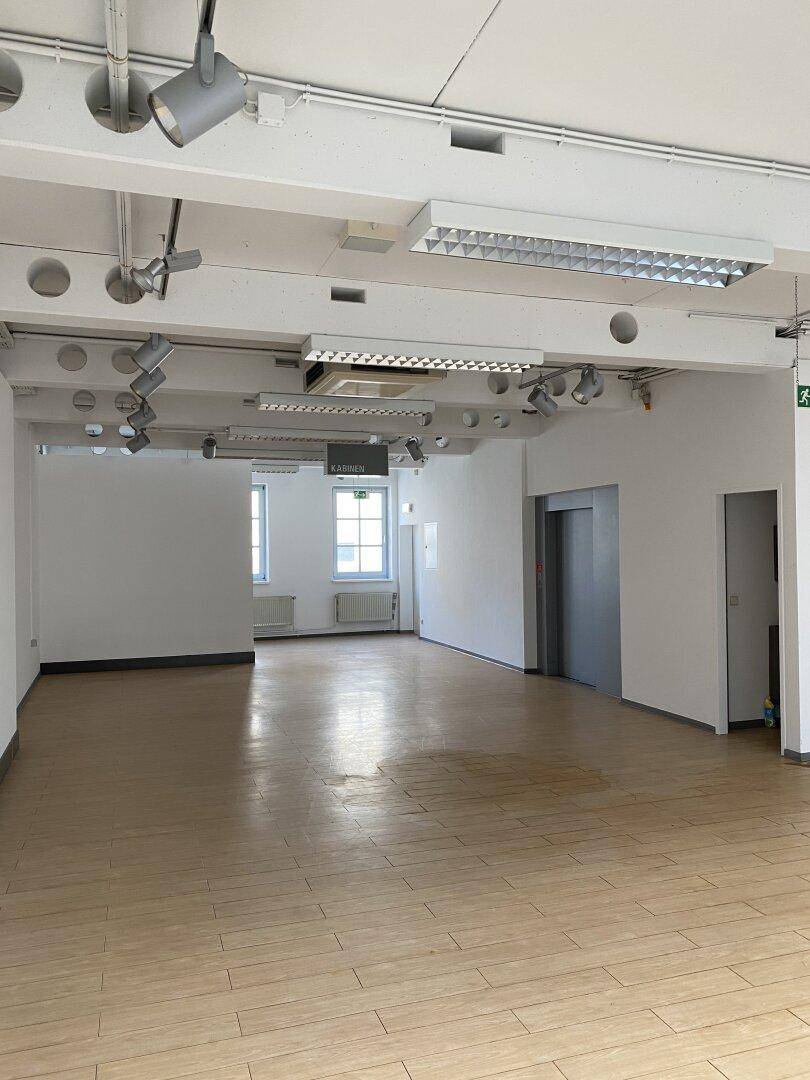 Großer, leerer Ladenbereich mit Holzboden, weißen Wänden und sichtbaren Deckeninstallationen, inklusive Aufzug.