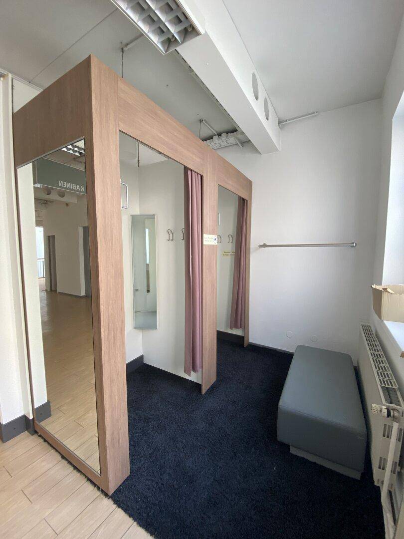 Umkleidekabinenbereich mit Spiegeln, Vorhängen, einer Sitzbank und dunklem Teppichboden.