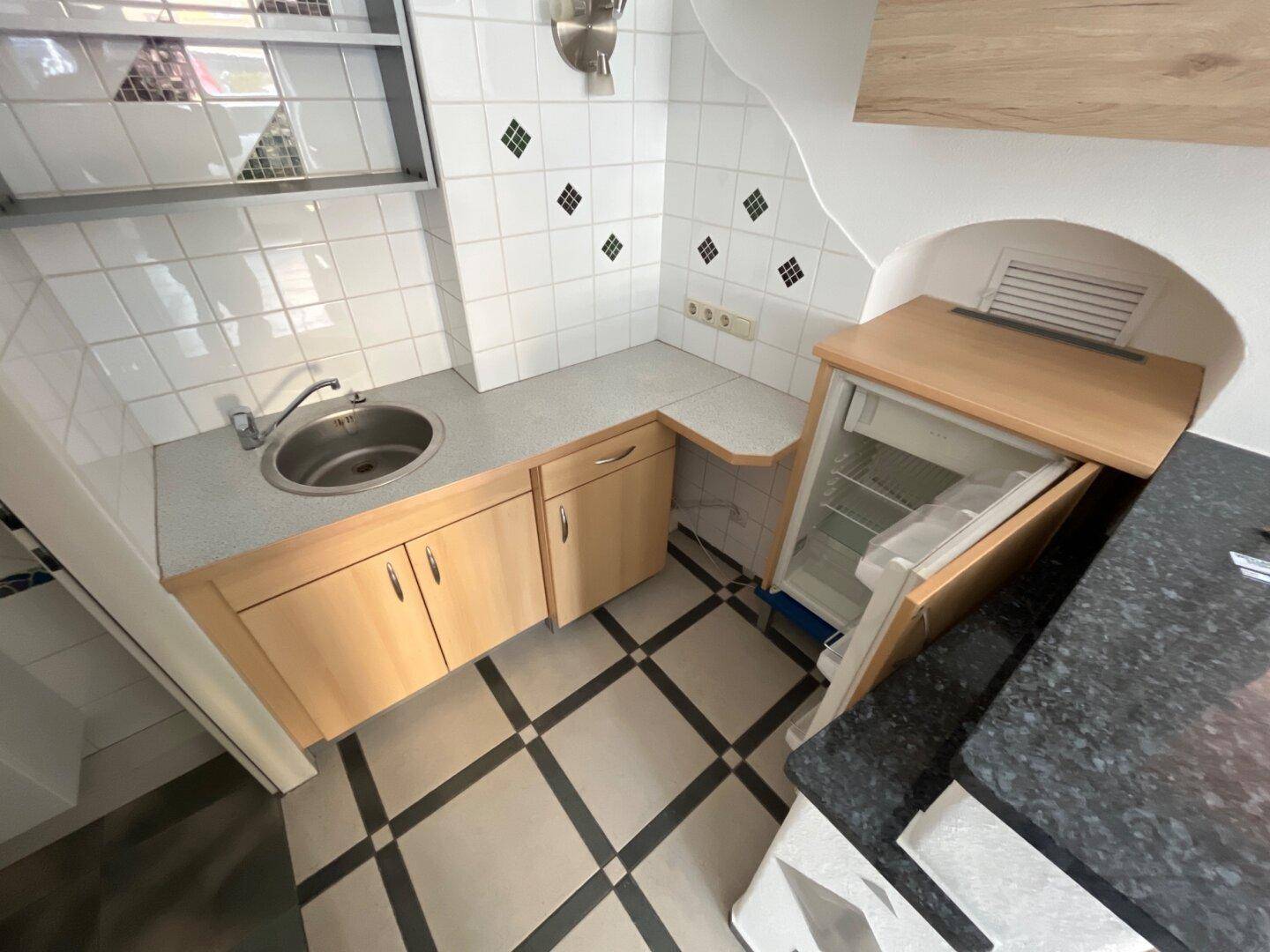 Kompakte Küchenzeile mit Spüle, Holzschränken und integriertem Kühlschrank, auf gemustertem Fliesenboden.