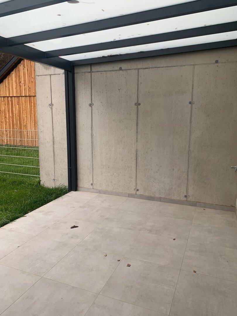 Überdachte Terrasse mit Fliesenboden und Betonwänden, angrenzend an eine Rasenfläche.