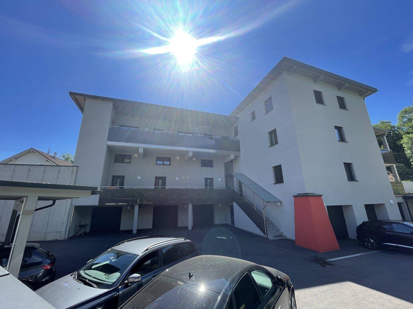 Rückansicht eines modernen Wohngebäudes mit Balkonen, Parkplätzen und Carport unter strahlender Sonne.