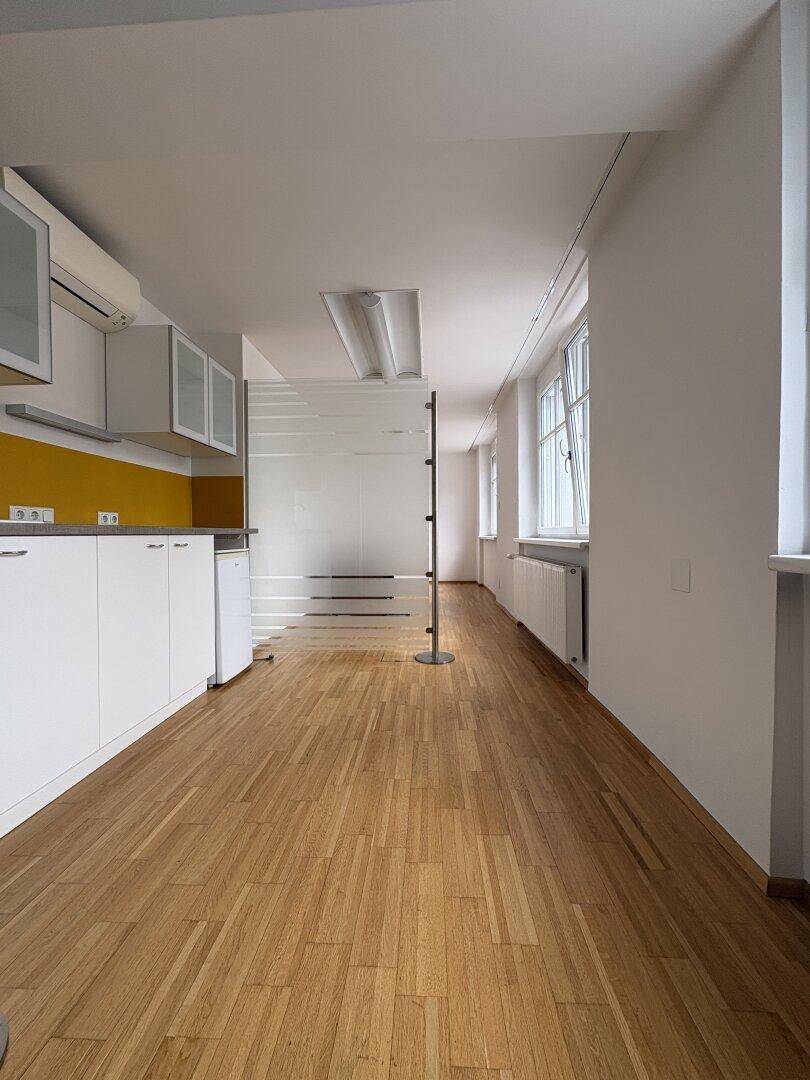 Langer Raum mit Parkettboden und einer modernen Küchenzeile mit weißen Schränken und gelber Rückwand.