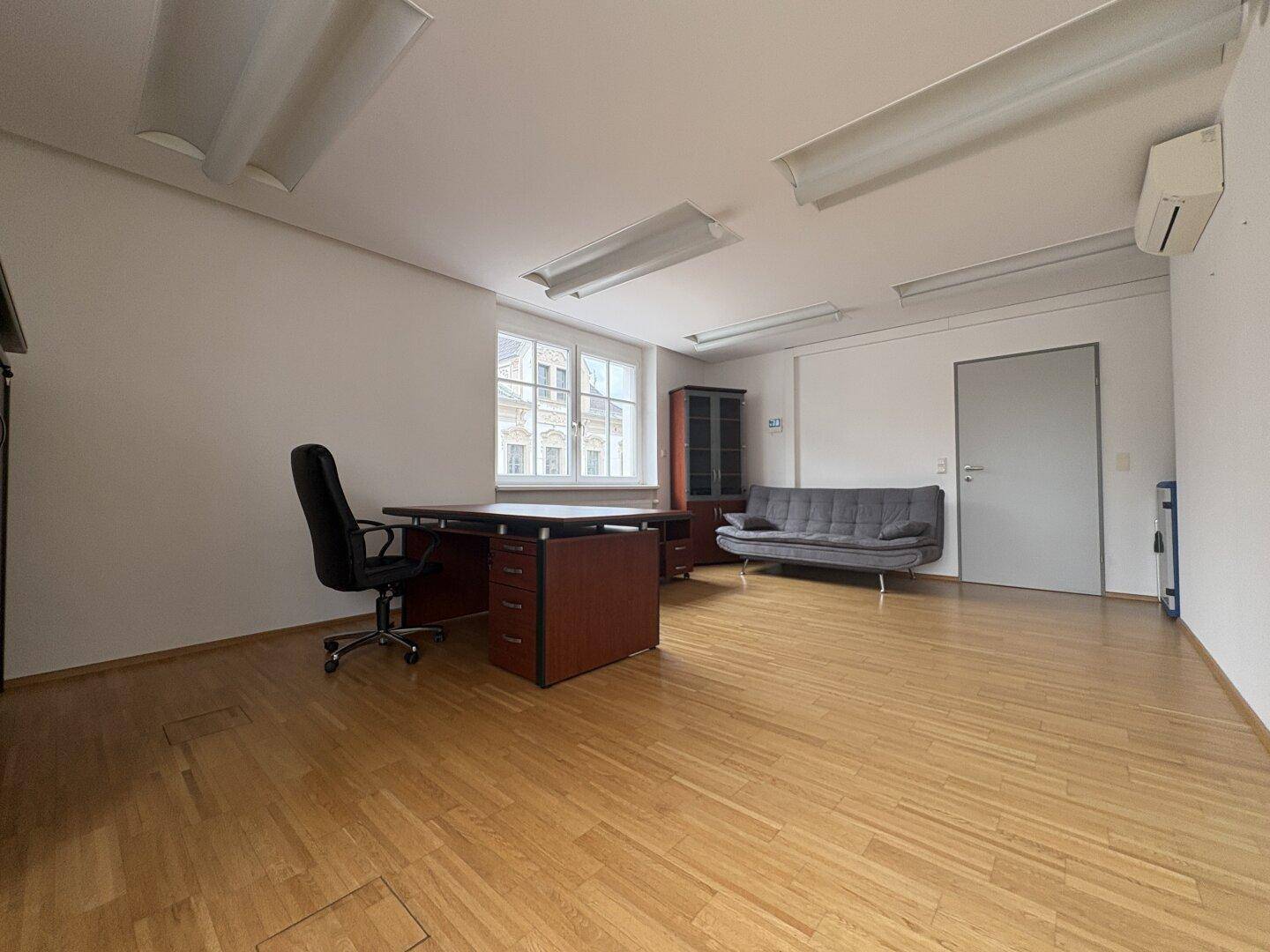 Geräumiges Büro mit Parkettboden, Schreibtisch, Sofa und Klimaanlage, ideal für produktives Arbeiten.