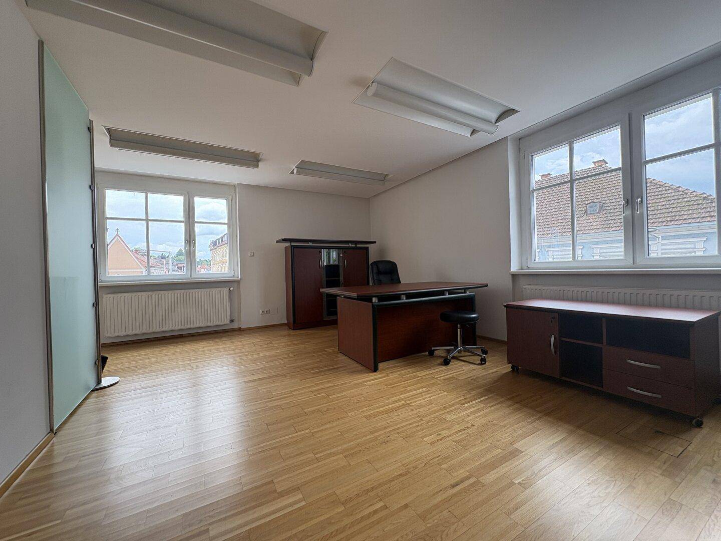 Büro mit zwei Fenstern, Parkettboden und einem Schreibtisch mit Bürostuhl, bereit zur Nutzung.