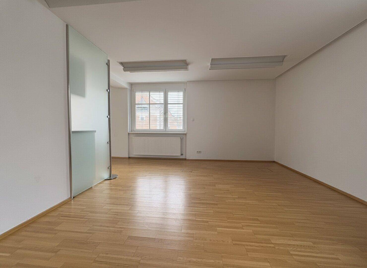 Helles Büro mit Parkettboden, weißen Wänden und einer Milchglaswand als Raumteiler.