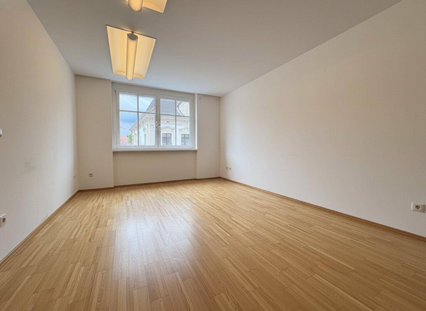 Leeres Büro mit hellem Parkettboden, weißen Wänden und einem großen Fenster, das viel Tageslicht bietet.