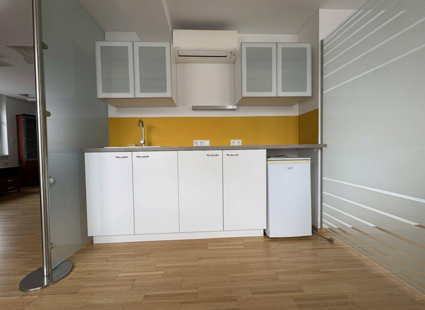 Moderne Küchenzeile mit weißen Schränken, gelber Rückwand und integriertem Kühlschrank in einem hellen Raum.