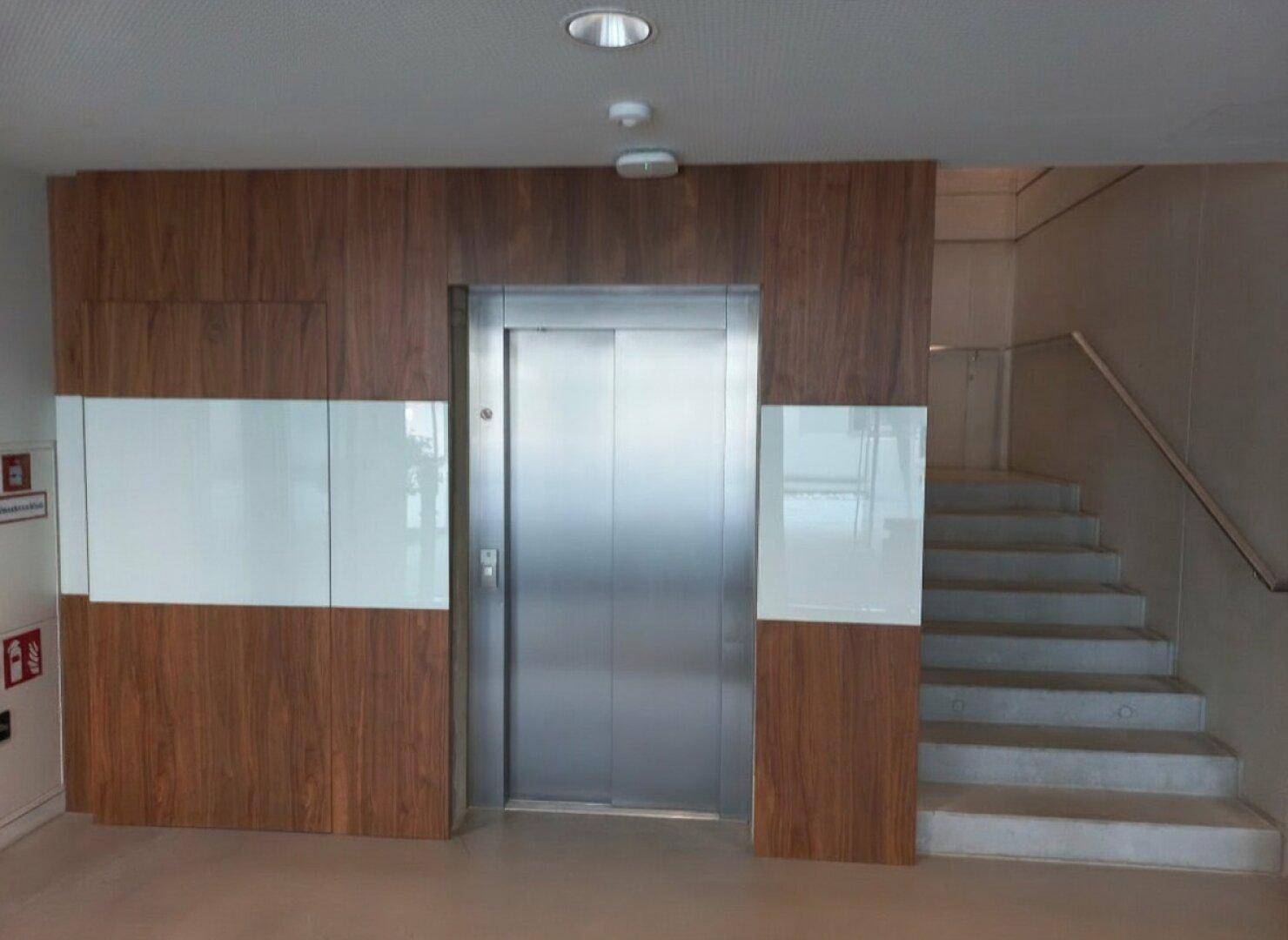 Moderner Eingangsbereich mit Aufzug und Treppe, verkleidet mit Holzpaneelen und weißen Akzenten.