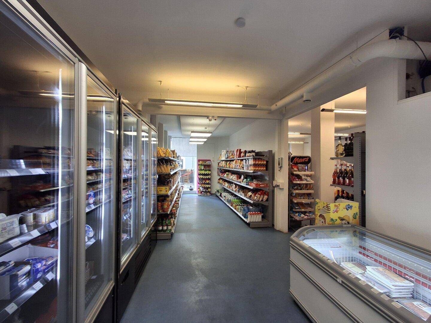 Gang in einem Mini-Markt mit mehreren Kühlvitrinen auf der linken Seite und Regalen auf der rechten Seite.