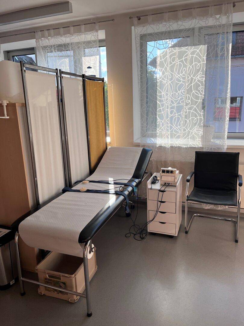 Detailansicht der Untersuchungsliege und der hellen Fensterfront mit Vorhängen.