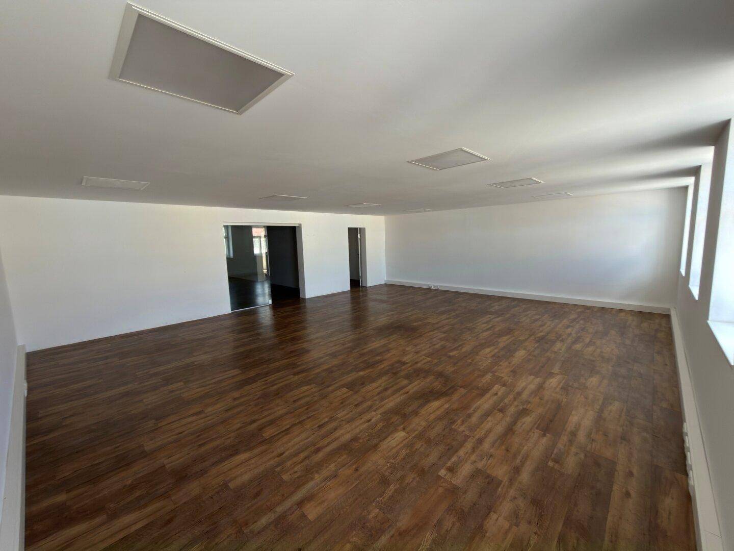 Heller, leerer Raum mit Holzboden und Zugang zu weiteren Bereichen, ausgestattet mit modernen Deckenleuchten.
