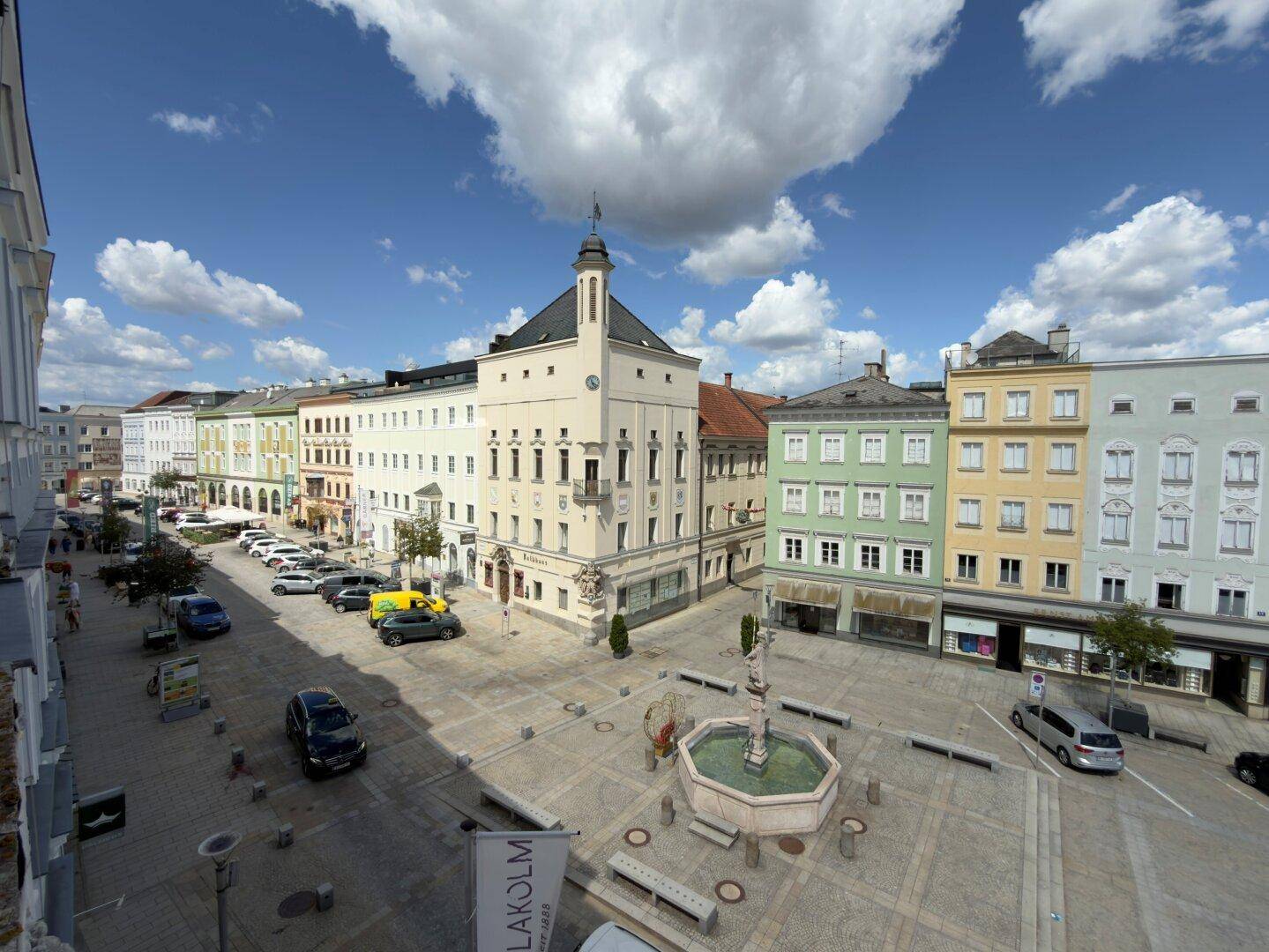 Panoramablick auf einen belebten Stadtplatz mit historischen Gebäuden, parkenden Autos und einem zentralen Brunnen.
