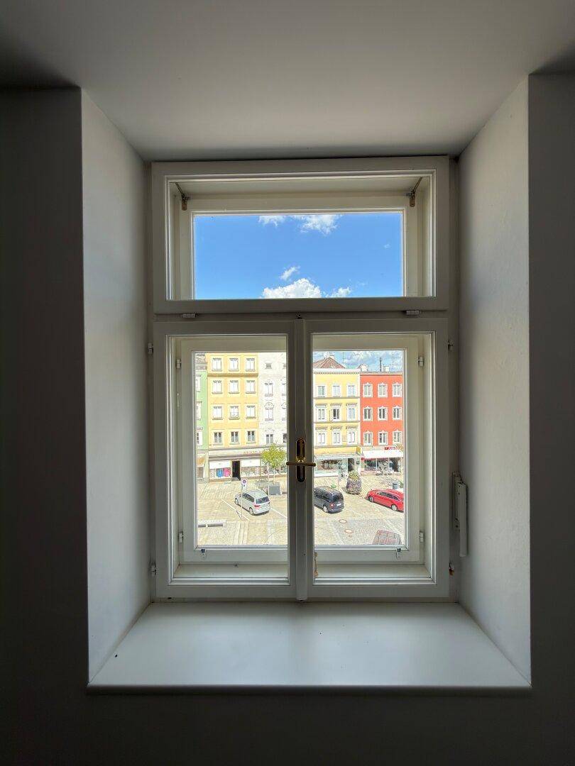 Blick aus einem Fenster auf den belebten Stadtplatz mit parkenden Autos und historischen Gebäuden unter blauem Himmel.