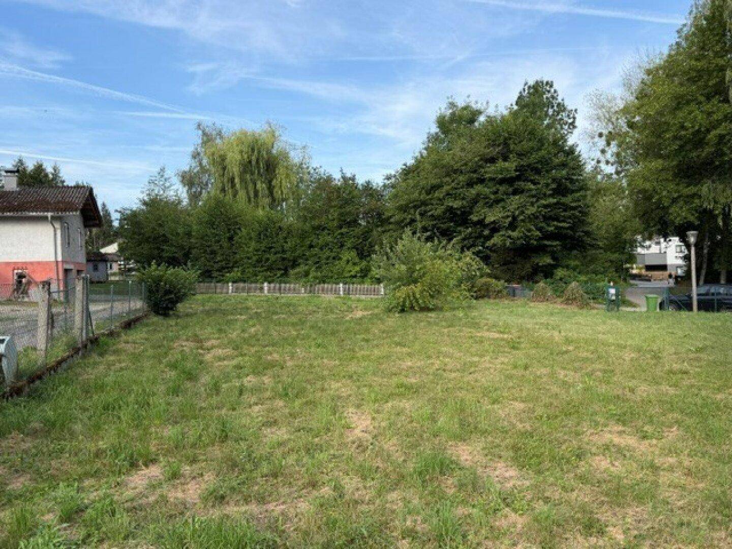 Weitläufige Rasenfläche mit einem Wohngebäude am Rand und dichtem Baumbestand im Hintergrund.