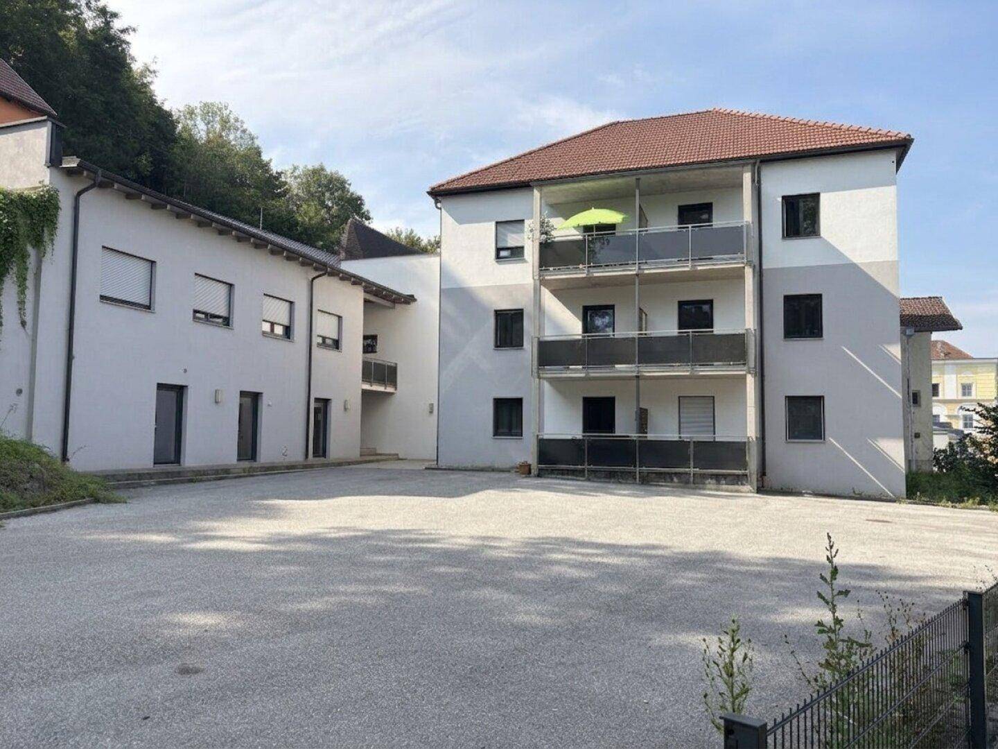 Mehrfamilienhaus mit Balkonen und großem Vorplatz, umgeben von Bäumen unter blauem Himmel.