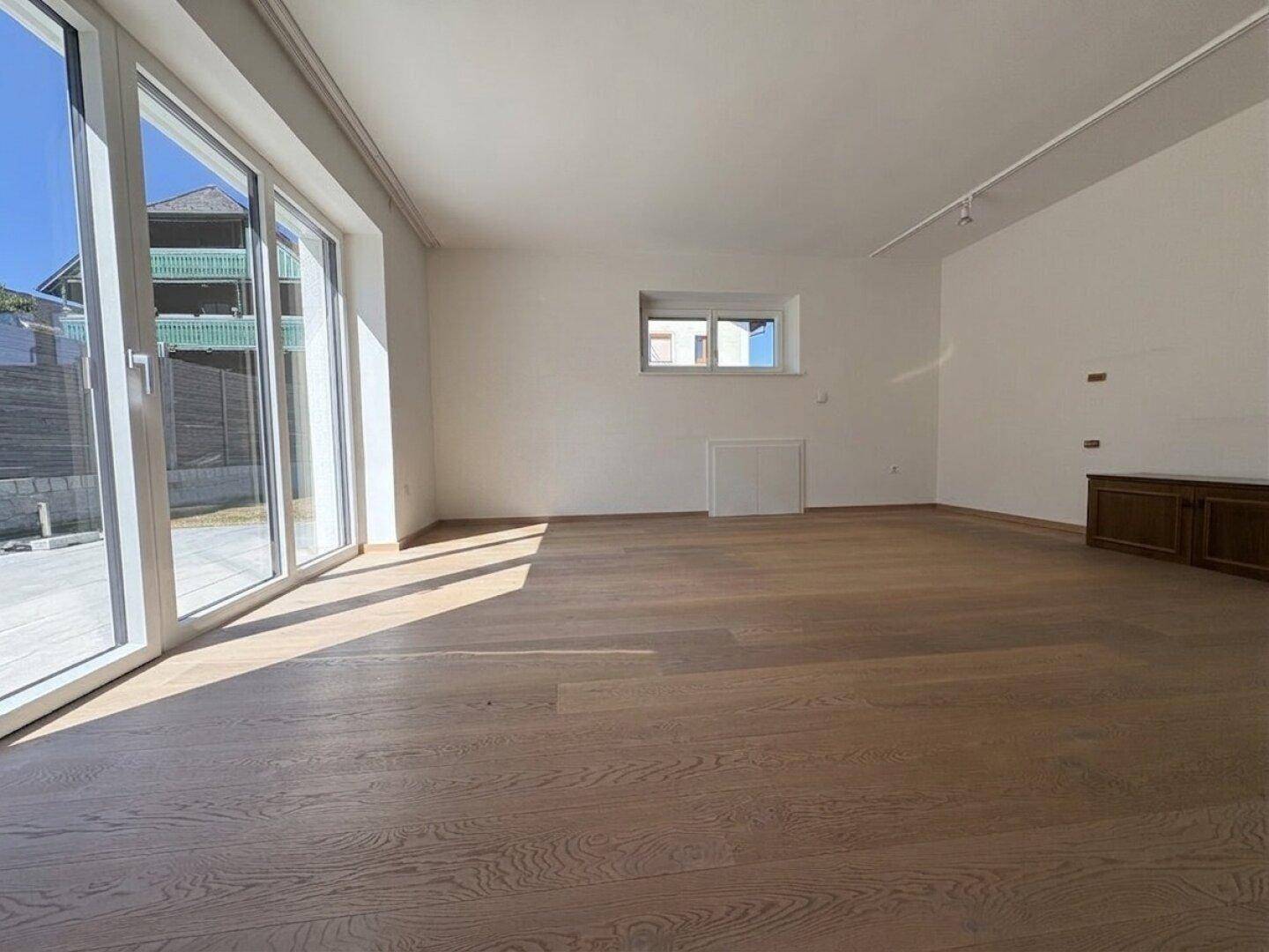 Lichtdurchflutetes Wohnzimmer mit Holzboden und großen Fenstern, die den Blick nach draußen freigeben.