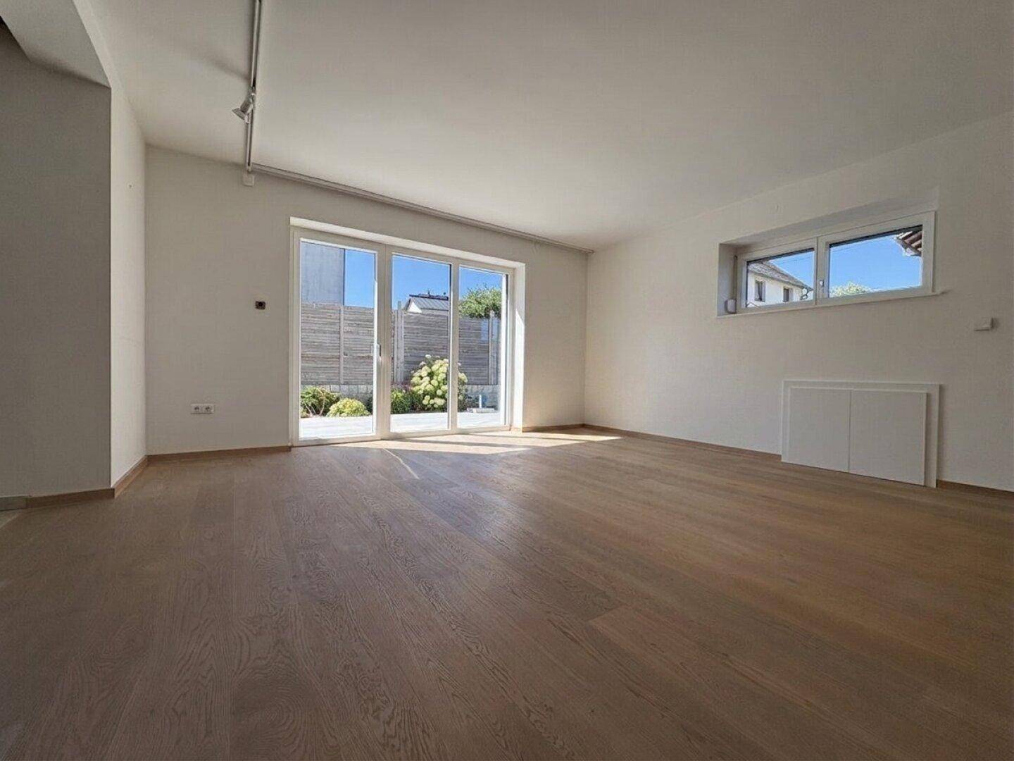 Geräumiges Wohnzimmer mit Holzboden und großen Fenstern, die viel Tageslicht hereinlassen.