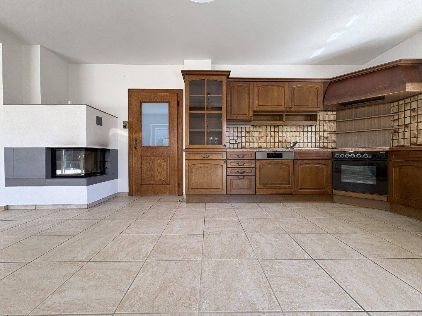 Geräumige Küche mit Holzeinbauten, Fliesenboden und einem modernen Kamin im Hintergrund des Raumes.