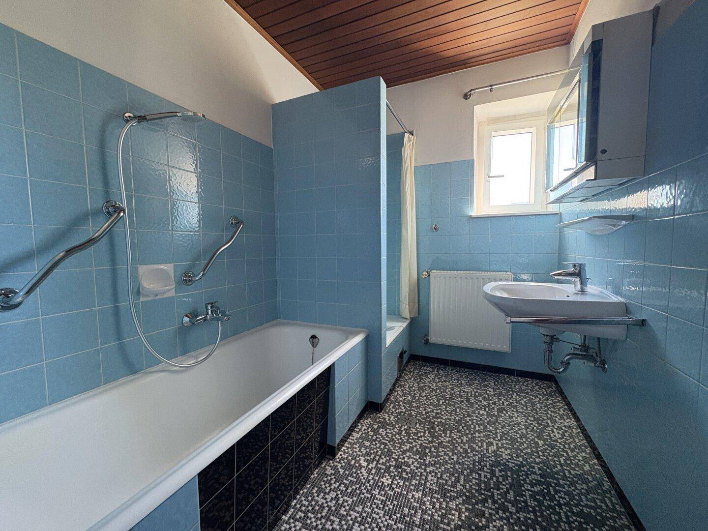 Badezimmer im Stil der 80er Jahre mit blauen Fliesen, Badewanne, Haltegriffen und Holzdecke.