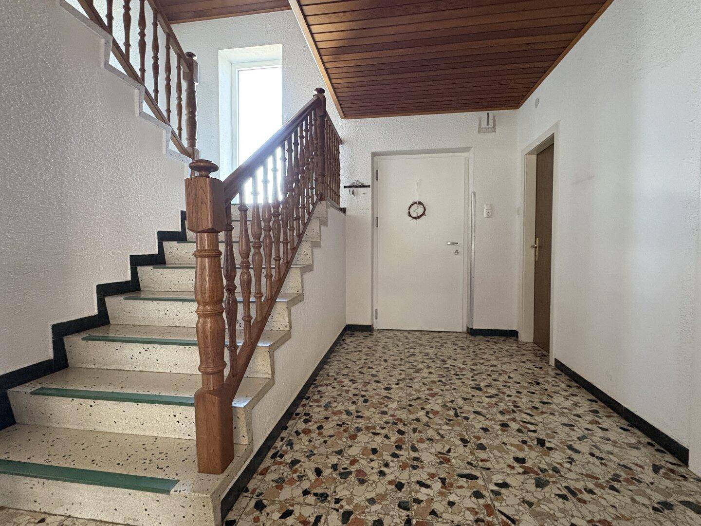 Heller Eingangsbereich mit Holztreppe, Terrazzoboden und Zugang zu verschiedenen Räumen.