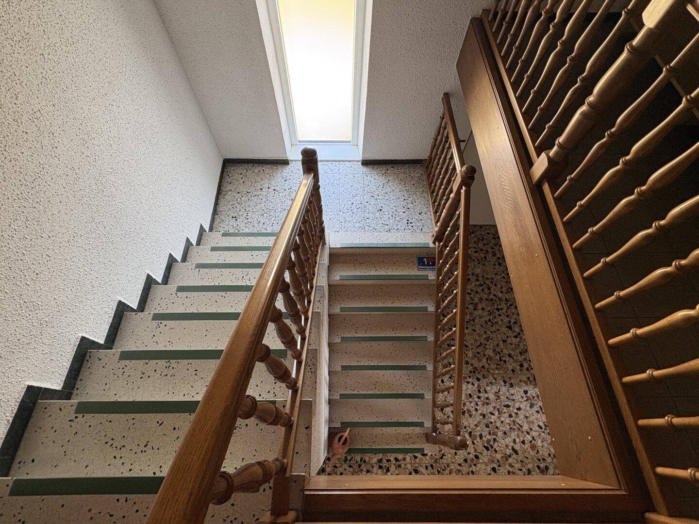 Treppenhaus mit Terrazzoboden, Holzgeländer und einem Oberlicht, das natürliches Licht spendet.