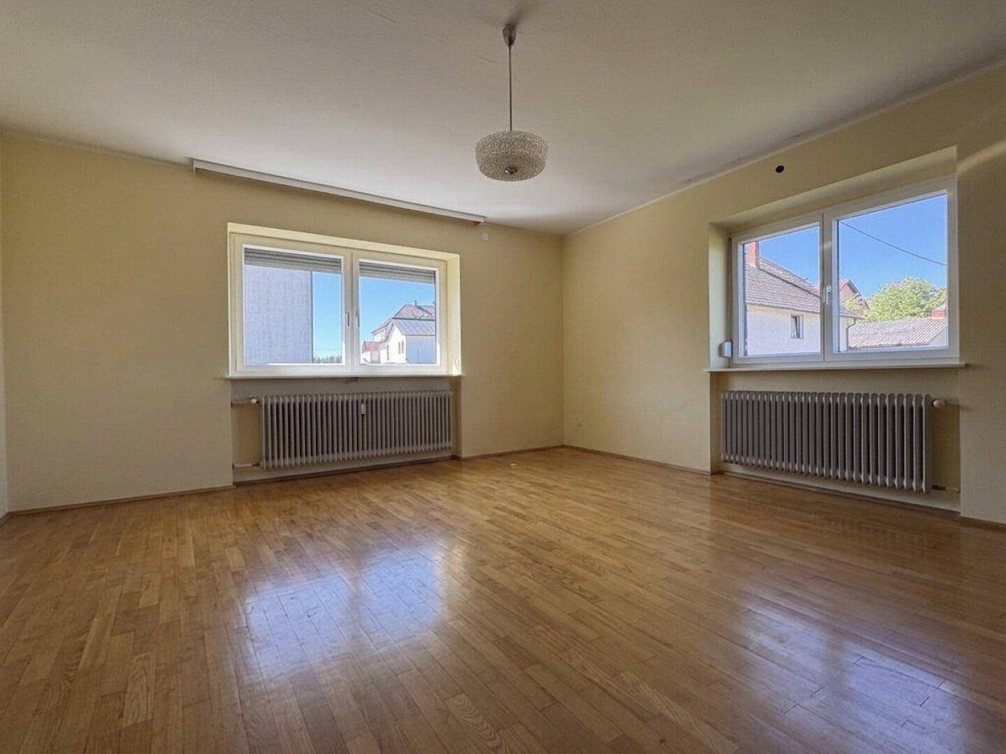 Helles Zimmer mit Parkettboden, zwei Fenstern und Heizkörpern unter den Fensterbänken.