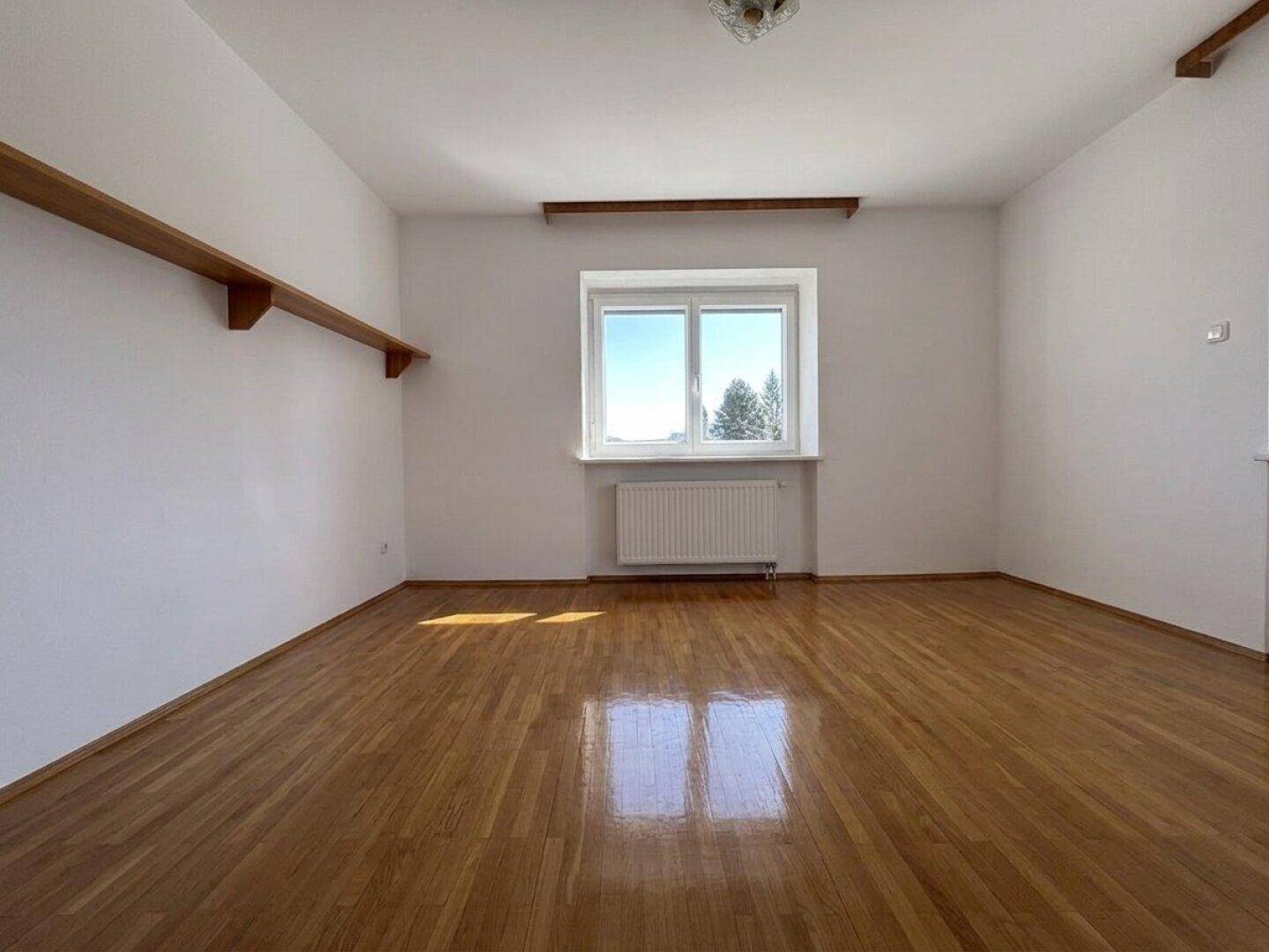 Helles Zimmer mit Parkettboden, einem Fenster und einer schlichten Wandgestaltung, ideal als Schlafzimmer.