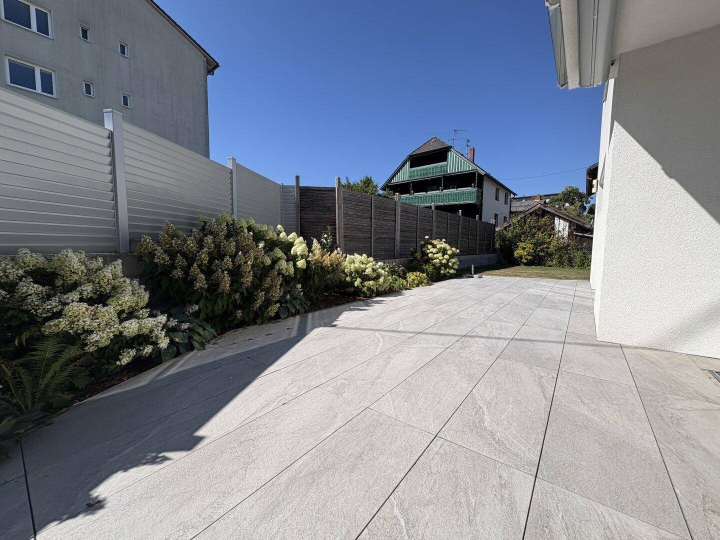 Große gepflasterte Terrasse mit Blumenbeeten und einem modernen Sichtschutzzaun.